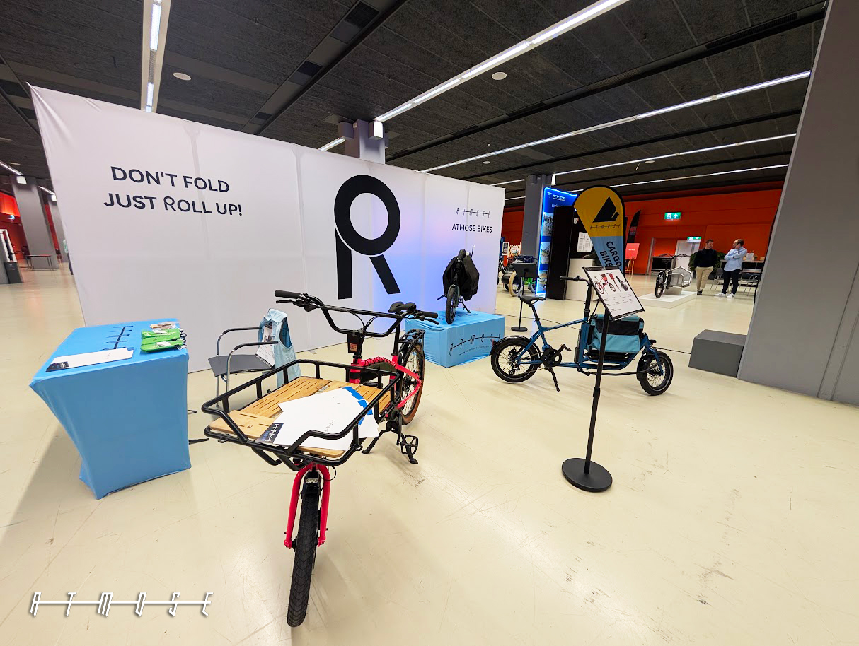 ATMOSE stand at ICBF 2026 in Utrecht, with a folding cargo bike in front of a white backdrop reading 'Don't fold, just roll up' and a large letter R