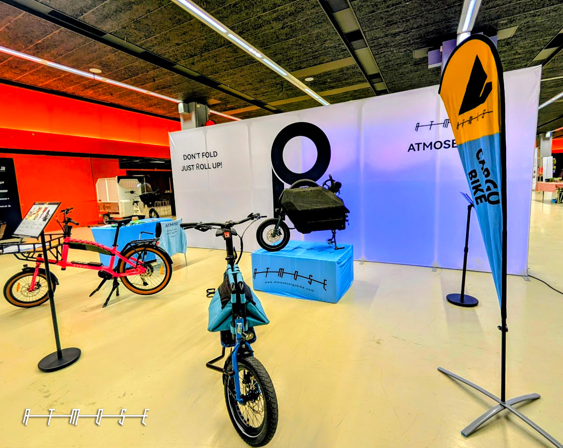 Wide view of the ATMOSE exhibition stand at ICBF 2026 in Utrecht, with the Model Q urban cargo bike on the right of the frame alongside other ATMOSE cargo bikes and branded flag banners