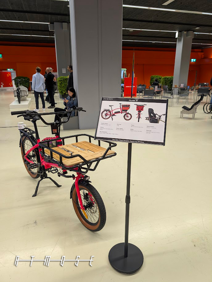 A pink ATMOSE cargo bike with a wooden front deck beside a spec board headed 